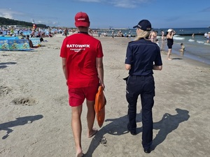 policjantka i ratownik WOPR idą brzegiem morza na plaży obleganej przez plażowiczów