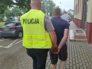 na zewnątrz komendy policjant prowadzi w kierunku radiowozu mężczyznę, który ma ręce skute za plecami kajdankami