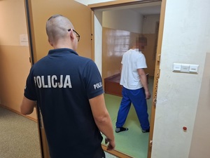 w celi stoi zatrzymany mężczyzna. Za nim, przy otwartych drzwiach celi stoi policjant służby dyżurnej