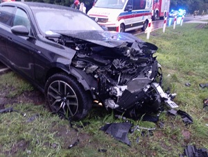 częściowo na jezdni i częściowo na trawie znajduje się czarne osobowe bmw z wgniecionym przodem. Za nim stoi karetka pogotowia, za karetką dwa policyjne radiowozy z włączonymi sygnałami błyskowymi