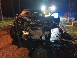 pora nocna. Na jezdni stoi czarne osobowe auto z wgniecionym przodem. Za nim znajduje się laweta i pracownik firmy holowniczej
