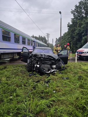przed torami na których znajduje się pociąg stoi czarne osobowe bmw z wgniecionym przodem. Za autem stoją strażacy, obok nich znajduje się karetka pogotowia