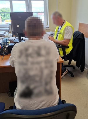 przy biurku siedzi policjant, który przesłuchuje siedzącego na krześle mężczyznę