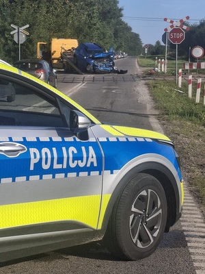 miejsce wypadku drogowego. Przed niestrzeżonym przejazdem kolejowym w poprzek jezdni stoi policyjny oznakowany radiowóz. Przed nim, za przejazdem kolejowym uszkodzone osobowe auto jest wciągane na lawetę