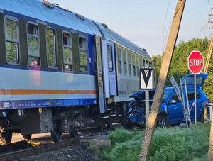 miejsce wypadku na przejeździe kolejowym. Na torach stoi pociąg. Obok znajduje się niebieski osobowy samochód z rozbitym przodem