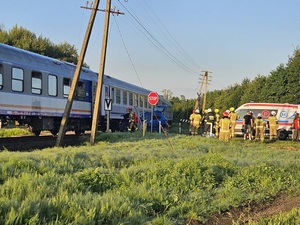 miejsce wypadku na przejeździe kolejowym. Na torach stoi pociąg. Obok znajduje się niebieski osobowy samochód z rozbitym przodem. Dalej stoją strażacy i karetka pogotowia