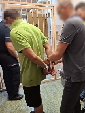 w korytarzu pomieszczenia dla osób zatrzymanych policjant zakłada mężczyźnie kajdanki na ręce trzymane z tyłu