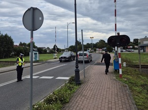 strzeżony przejazd kolejowy. Policjantka ruchu drogowego kieruje ruchem pojazdów. Pracownik kolei stoi przed przejazdem
