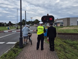 strzeżony przejazd kolejowy. Przed opuszczonymi rogatkami stoi rowerzysta, obok policjantka i pracownik kolei