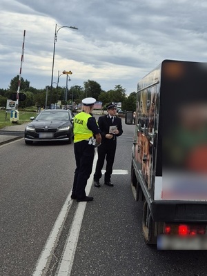strzeżony przejazd kolejowy. Przed przejazdem stoi samochód dostawczy przy którym stoją policjantka ruchu drogowego i pracownik kolei