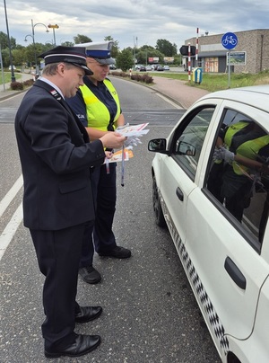 strzeżony przejazd kolejowy. Przed przejazdem stoi samochód osobowy przy którym stoją policjantka ruchu drogowego i pracownik kolei. Kolejarz trzyma w rękach ulotki i opaski odblaskowe