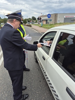 strzeżony przejazd kolejowy. Przed przejazdem stoi samochód osobowy przy którym stoją policjantka ruchu drogowego i pracownik kolei. Kolejarz przekazuje kierowcy ulotki i opaskę odblaskową