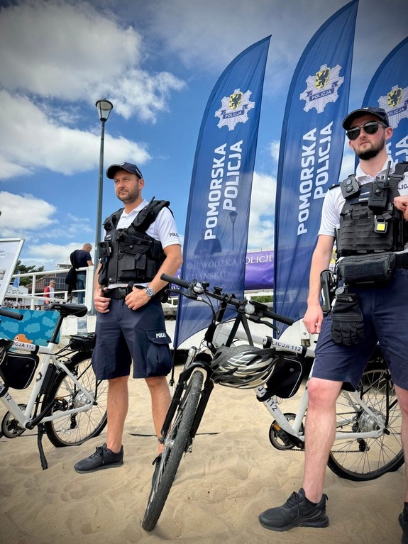 na plaży przy rowerach stoją dwaj policjanci służby patrolowej. Za nimi są powiewające flagi z napisem Pomorska Policja