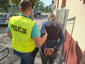 policjant w kamizelce odblaskowej z napisem policja prowadzi na zewnątrz komendy policji mężczyznę, który ma ręce skute za plecami kajdankami