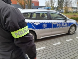 policjant w cywilnym ubraniu i z opaską z napisem policja, umieszczoną na rękawie kurtki, stojący przed oznakowanym policyjnym radiowozem