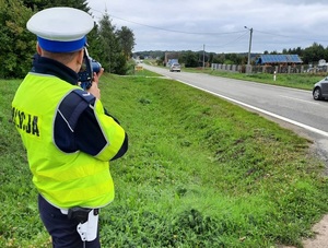 policjant ruchu drogowego, stojący przy drodze i mierzący prędkość przejeżdżających pojazdów za pomocą ręcznego miernika prędkości