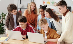 w klasie szkolnej dwoje uczniów siedzi w ławce przed laptopami. Za nimi stoją inni uczniowie