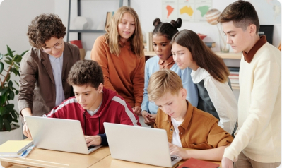 w klasie szkolnej dwoje uczniów siedzi w ławce przed laptopami. Za nimi stoją inni uczniowie