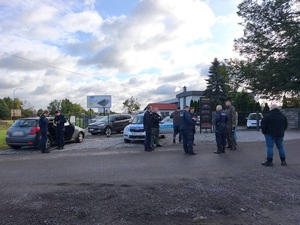 przed komisem samochodowym stoi policyjny radiowóz w pobliżu którego znajduje się kilkoro policjantów w umundurowaniu służbowym i w cywilnych ubraniach