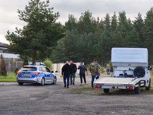 grupa nieumundurowanych policjantów kieruje się w stronę lasu. Obok stoi policyjny oznakowany radiowóz