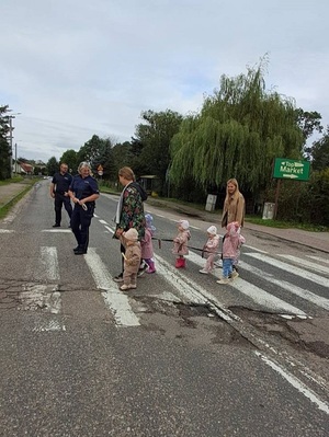 przez pasy przechodzą małe dzieci w towarzystwie dwóch opiekunek. Na środku drogi stoi policjant, przy pasach znajduje się policjantka