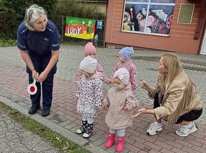 przed przejściem dla pieszych stoi czworo małych dzieci. Przy nich klęczy opiekunka. Obok stoi pochylona w stronę dzieci policjantka trzymająca tarczę do zatrzymywania pojazdów