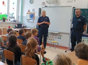 w klasie szkolnej na tle tablicy stoi dwoje policjantów. Przed nimi w ławkach siedzą uczniowie