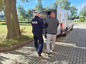policjant prowadzi w kierunku nieoznakowanego radiowozu mężczyznę, który ma ręce i nogi skute kajdankami
