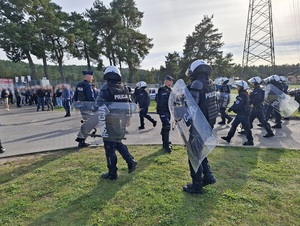 kordon policjantów ubranych w sprzęt przeciwuderzeniowy, zmierzających w stronę wejścia na stadion, przed którym stoją pseudokibice