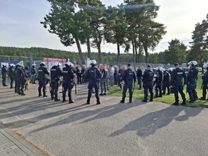 policjanci ubrani w sprzęt przeciwuderzeniowy, otaczający grupę pseudokibiców stojących przed wejściem na stadion