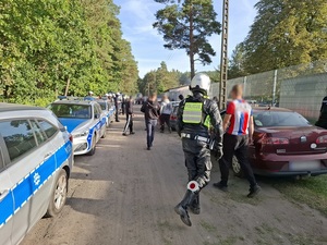 pseudokibice zmierzający w stronę klatki dla kibiców przyjezdnych na stadionie. Wzdłuż drogi są zaparkowane policyjne radiowozy przy których stoją policjanci ubrani w sprzęt przeciwuderzeniowy. Za pseudokibicami znajduje się policjant ruchu drogowego ubrany w kombinezon motocyklisty