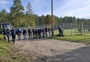 kordon policjantów ubranych w sprzęt przeciwuderzeniowy, z tarczami, stojący na zewnątrz stadionu, za sektorem kibiców przyjezdnych. Za nimi jest zaparkowany autokar