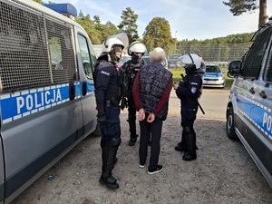 przed stadionem, pomiędzy dwoma radiowozami stoi mężczyzna, który ma ręce skute za placami kajdankami. Przy nim stoi troje policjantów ubranych w sprzęt przeciwuderzeniowy