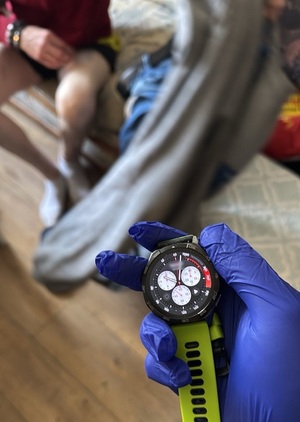 na pierwszym planie znajduje się dłoń w lateksowej rękawiczce trzymająca smartwatch. W tle na kanapie siedzi mężczyzna