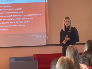 w klasie szkolnej przed siedzącymi w ławkach uczniami stoi policjantka. Za nią na tablicy wyświetla się prezentacja