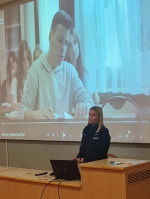 w szkolnej auli przy stole siedzi policjantka. Za nią na ścianie jest wyświetlony film