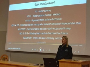w szkolnej auli przy stole siedzi policjantka. Za nią na ścianie wyświetlają się numery telefonów instytucji pomocowych