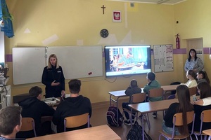 w klasie szkolnej przed siedzącymi w ławkach uczniami stoi policjantka. Za nią na tablicy wyświetla się film