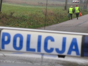 dwie osoby ubrane w kamizelki odblaskowe idą lewą stroną jezdni. Za nimi znajduje się policyjny radiowóz