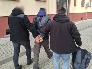 dwaj nieumundurowani policjanci kryminalni prowadzą w kierunku wejścia do budynku komendy mężczyznę, który ma ręce skute za plecami kajdankami