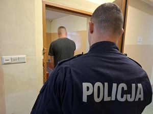 w celi stoi mężczyzna, odwrócony tyłem do obiektywu aparatu. Za nim, przy otwartych drzwiach celi stoi policjant służby dyżurnej