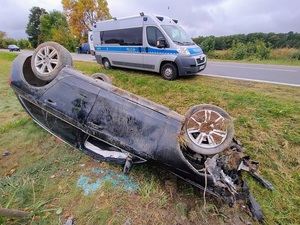 poza jezdnią, w rowie, znajduje się przewrócony na dach, osobowy samochód z licznymi uszkodzeniami. Za tym autem, na poboczu stoi policyjny radiowóz