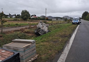 poza jezdnią, w rowie, znajduje się przewrócony na dach, osobowy samochód z licznymi uszkodzeniami. Przed nim, na poboczu są ustawione dwie palety z kostką brukową. Za tym autem, na poboczu stoi policyjny radiowóz