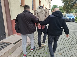 dwaj nieumundurowani policjanci kryminalni prowadzą w stronę wejścia do budynku komendy mężczyznę, który ma ręce skute za plecami kajdankami. Mężczyzna ma ubłocone ubranie, nie ma jednego buta
