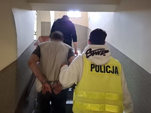 policjant w kamizelce odblaskowej z napisem &quot;policja&quot; prowadzi schodami do góry mężczyznę, który ma ręce skute za plecami kajdankami