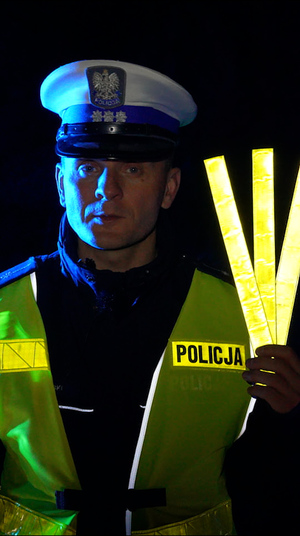 pora nocna. Na dworze znajduje się policjant ruchu drogowego, który trzyma w ręce trzy opaski odblaskowe