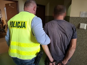 policjant w kamizelce odblaskowej z napisem policja prowadzi korytarzem mężczyznę, który ma ręce skute za plecami kajdankami