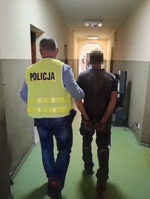 policjant w kamizelce odblaskowej z napisem policja prowadzi korytarzem mężczyznę, który ma ręce skute za plecami kajdankami