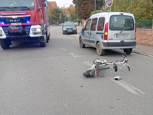 miejsce zdarzenia drogowego. Na środku jezdni leży hulajnoga elektryczna. Przed nią stoi szary osobowy renault z urwanym lusterkiem. Obok stoi wóz straży pożarnej