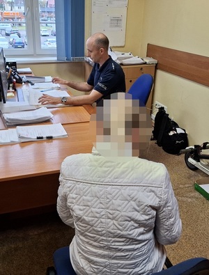 siedzący przed komputerem policjant przesłuchuje kobietę, która siedzi przy stoliku obok biurka policjanta